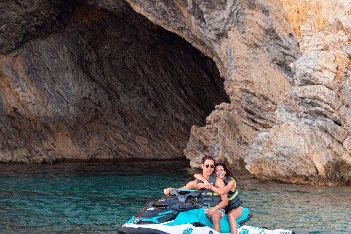 Two people on a jet ski near a rocky cave with turquoise water.