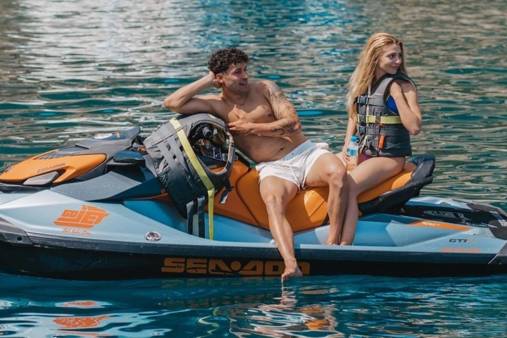 Two people on a jet ski in a sunny ocean, wearing life jackets and smiling.