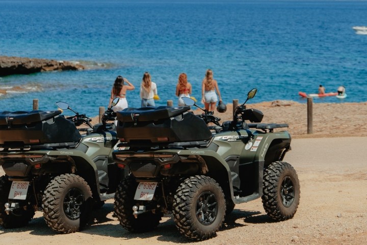 Two ATVs parked near a beach with people in the background and a boat on the water.