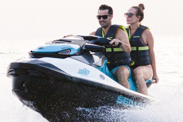 Two people riding a jet ski on the water, both wearing life vests and sunglasses.