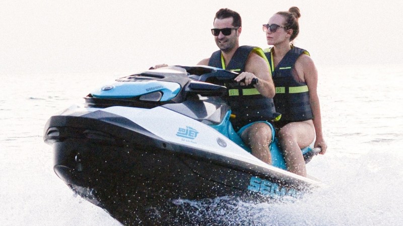 Two people riding a jet ski on the water, both wearing life vests and sunglasses.