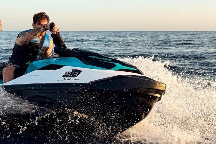 Person riding a jet ski on the ocean creating splash, with a rocky cliff in the background.