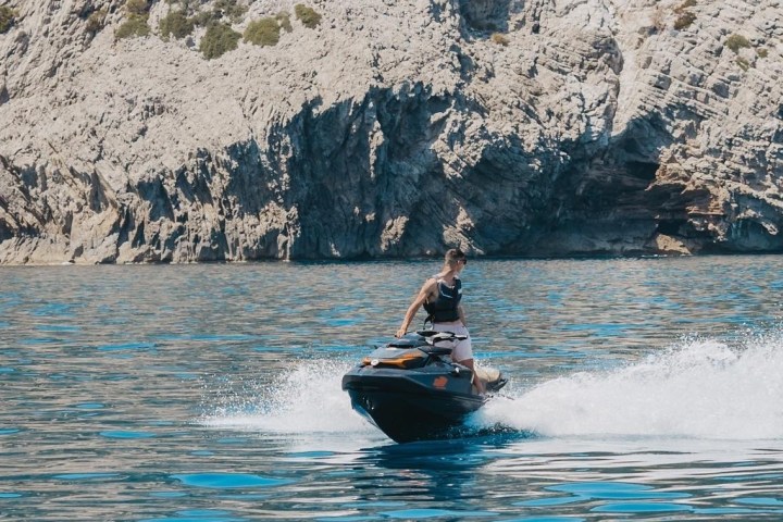 Person riding a jet ski on a calm sea with rocky cliffs in the background.