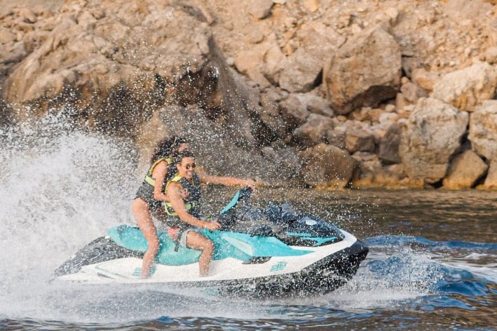 Two people on a jet ski creating splashes near a rocky coastline.
