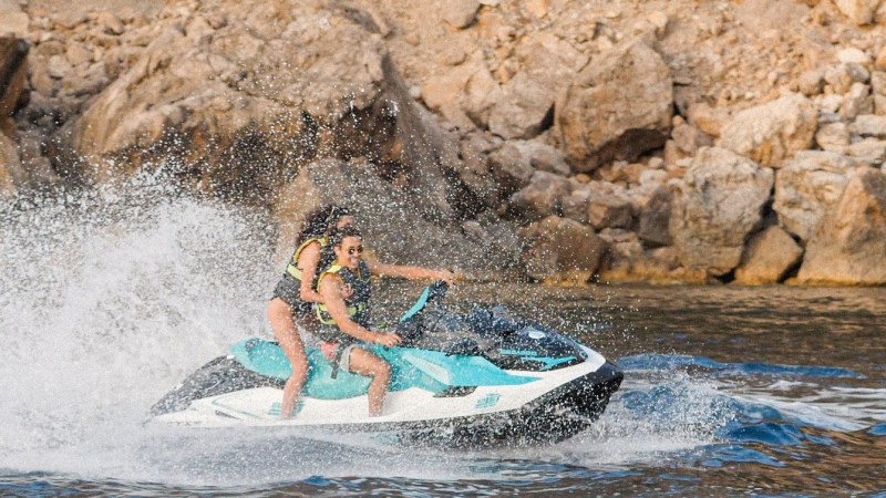 Two people on a jet ski creating splashes near a rocky coastline.