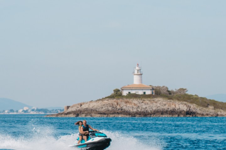 Two people on a jet ski near the ocean with a lighthouse on a distant island.