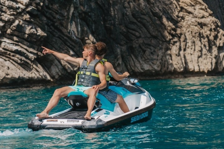 Two people on a jet ski pointing at rocks in blue ocean water.