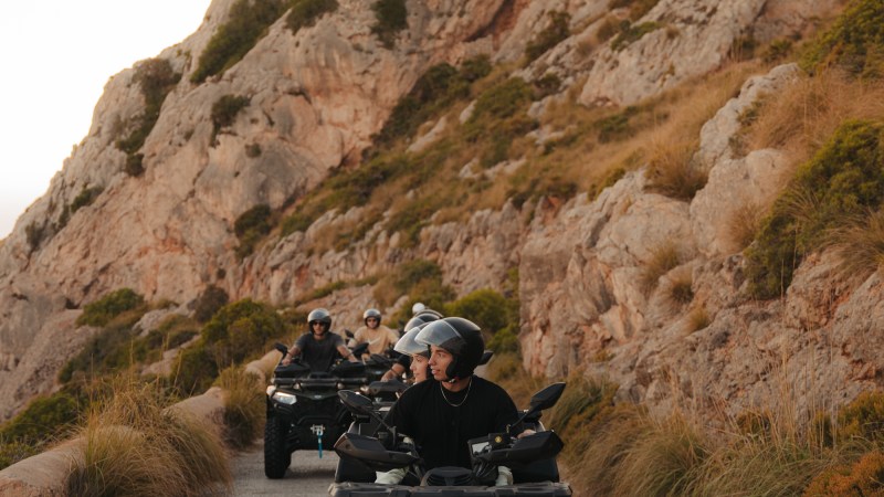 People riding ATVs on a narrow path beside rocky hills at sunset.