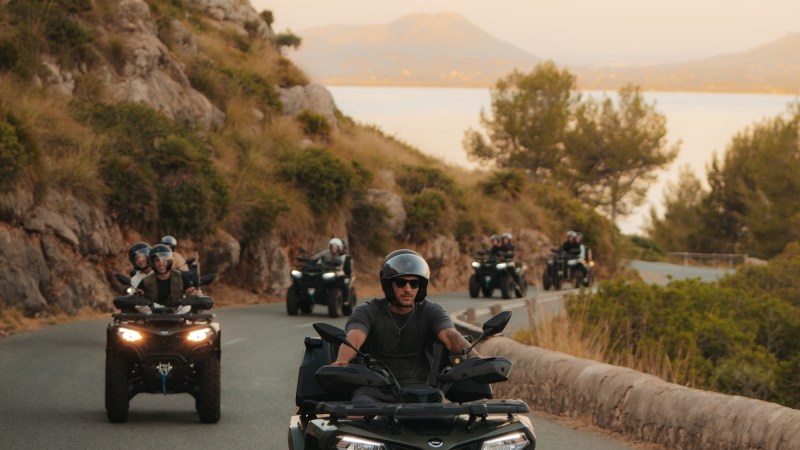 Group of people riding ATVs on a winding road with hills and distant mountains.
