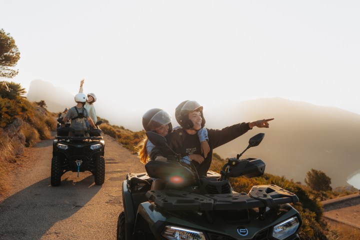 Two people on ATVs pointing at sunrise on a hilltop road.