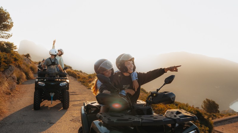 Two people on ATVs pointing at sunrise on a hilltop road.