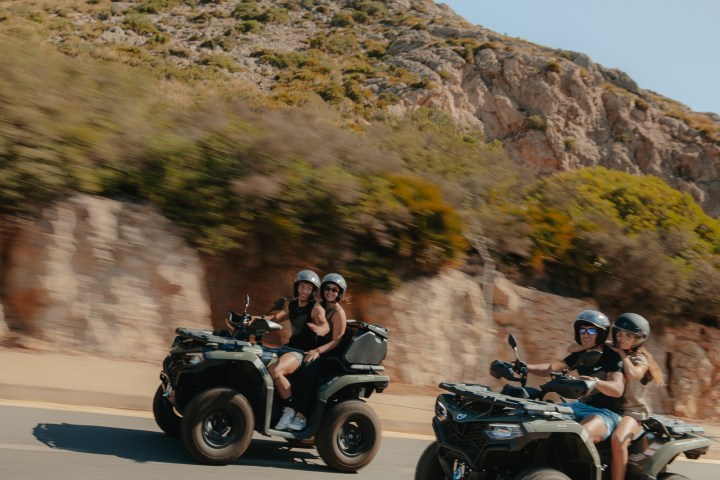 Two ATVs with riders wearing helmets, driving on a road against a rocky hill backdrop.