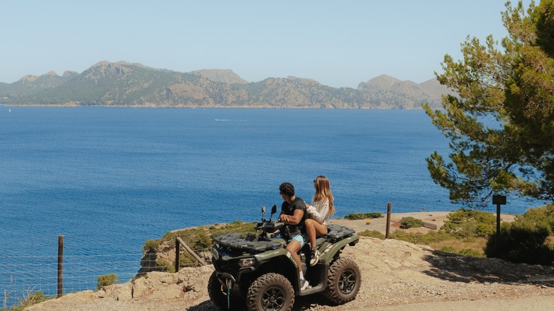 Two people on an ATV overlooking a blue sea with distant mountains.