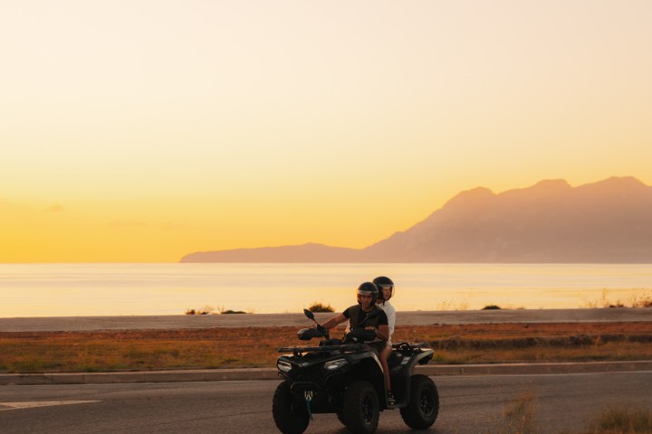 Two people on an ATV ride along a coastal road at sunset with mountains in the background.