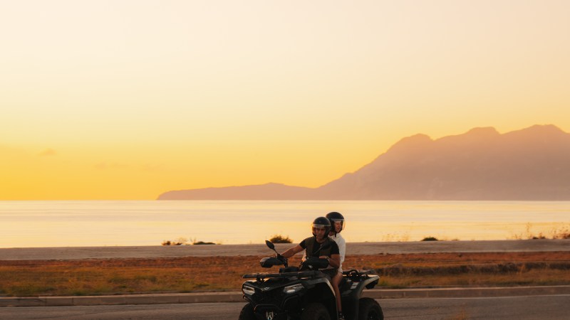 Two people on an ATV ride along a coastal road at sunset with mountains in the background.