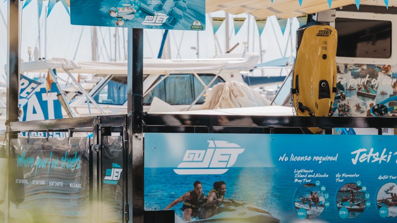 Jet ski rental booth with banners and photos, boats and marina in background.