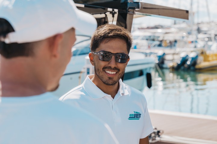 Two men in white shirts near a marina, one wearing sunglasses and smiling.