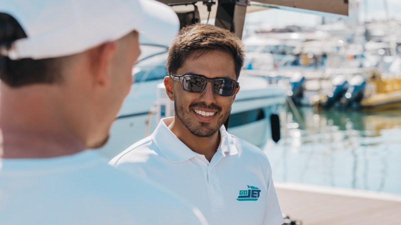 Two men in white shirts near a marina, one wearing sunglasses and smiling.