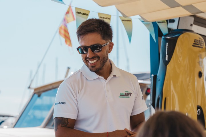 Man in sunglasses and white polo smiling under a canopy near boats.