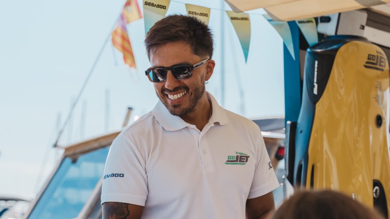 Man in sunglasses and white polo smiling under a canopy near boats.