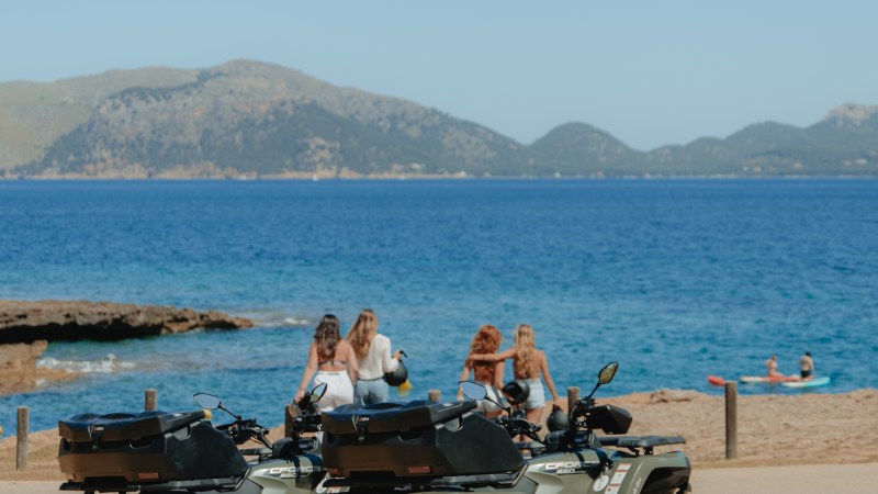 Two ATVs parked on a beach with people in the background and mountains across the water.