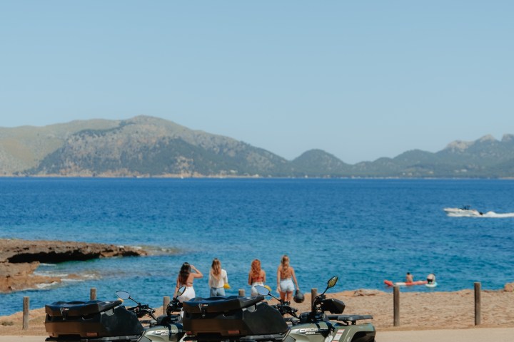 Two ATVs parked by a scenic beach with people and mountains in the background.