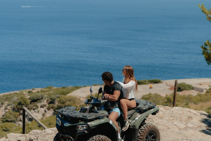 Two people on an ATV overlooking a coastal landscape with mountains and sea.