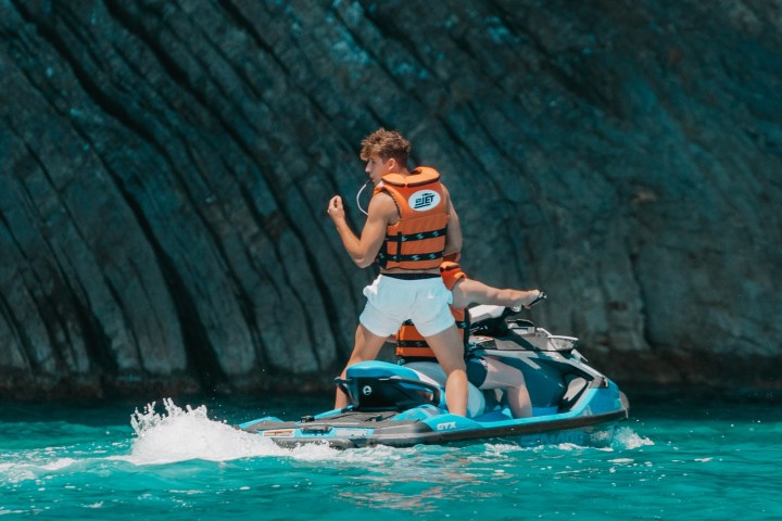 Person on a jet ski wearing a life jacket near a rocky cliff in turquoise water.
