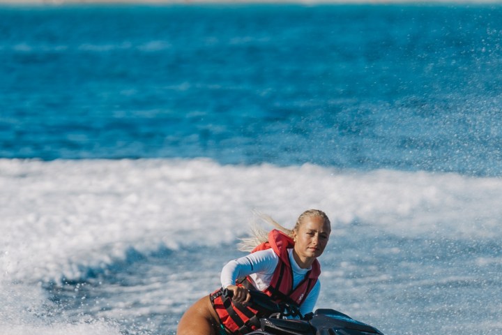 Person riding a jet ski on the ocean with a beach and buildings in the background.