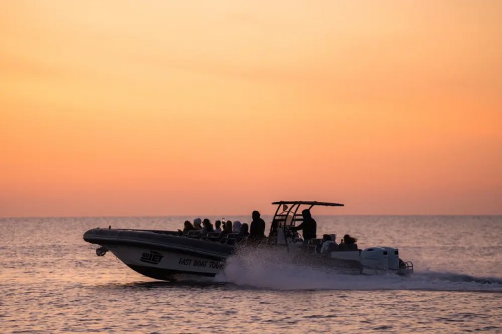 Silhouette of a speedboat with passengers against an orange sunset over the sea.