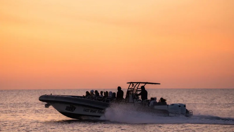 Silhouette of a speedboat with passengers against an orange sunset over the sea.