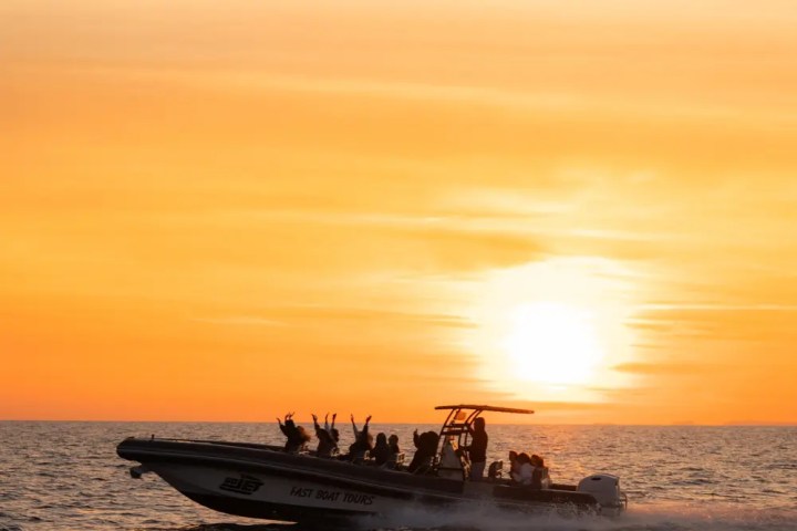 Silhouetted boat with passengers against an orange sunset on the sea.
