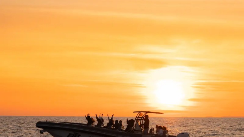 Silhouetted boat with passengers against an orange sunset on the sea.