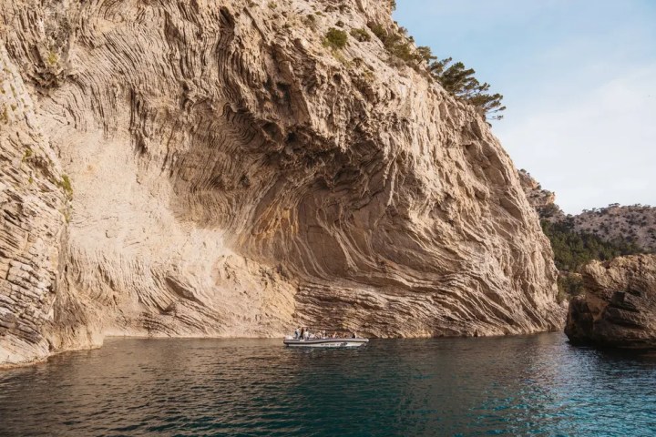 Small boat near large rocky cliff with layered patterns and trees.