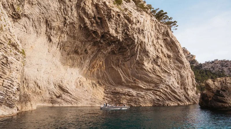 Small boat near large rocky cliff with layered patterns and trees.