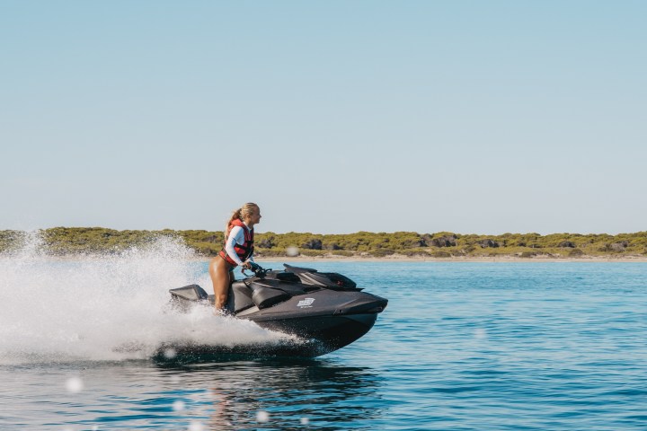 Person riding a jet ski on calm water with a distant shoreline under a clear sky.